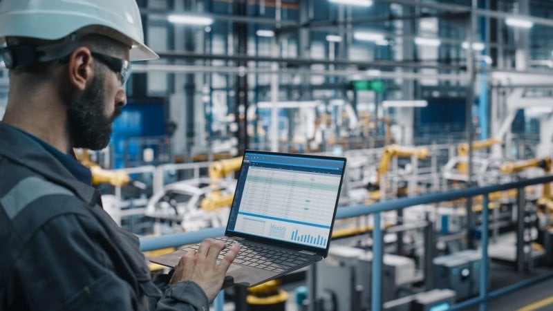 A quality engineer looking at a computer while he is standing on a factory floor.