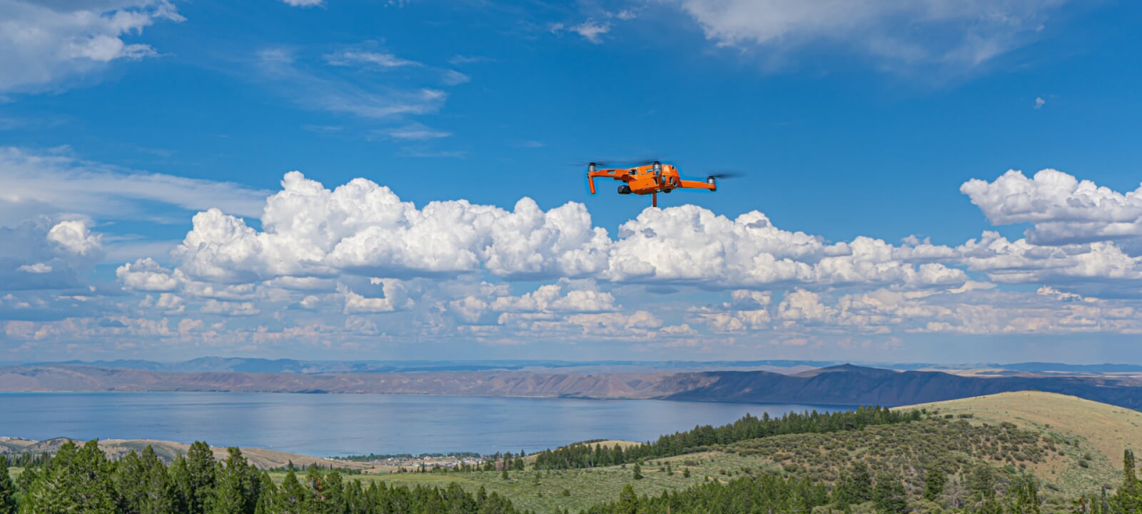 An orange drone flies over a field, a forest, and a body of water.