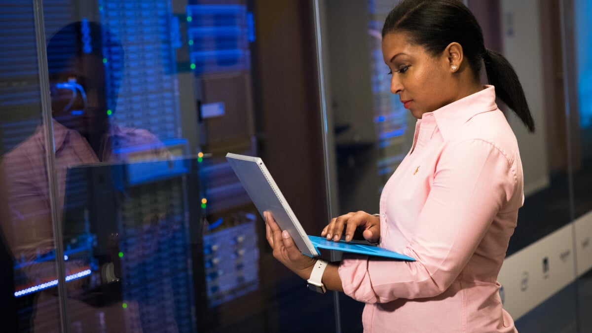 Young black cybersecurity professional stands in front of a computer mainframe while working on her laptop.
