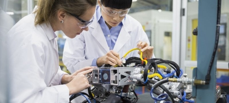 Two female engineers in lab coats working on a wired piece of equipment.