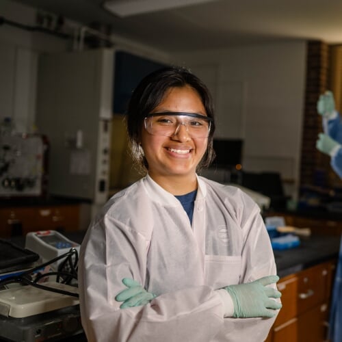 Graduate student in lab coat and safety glasses in a lab.