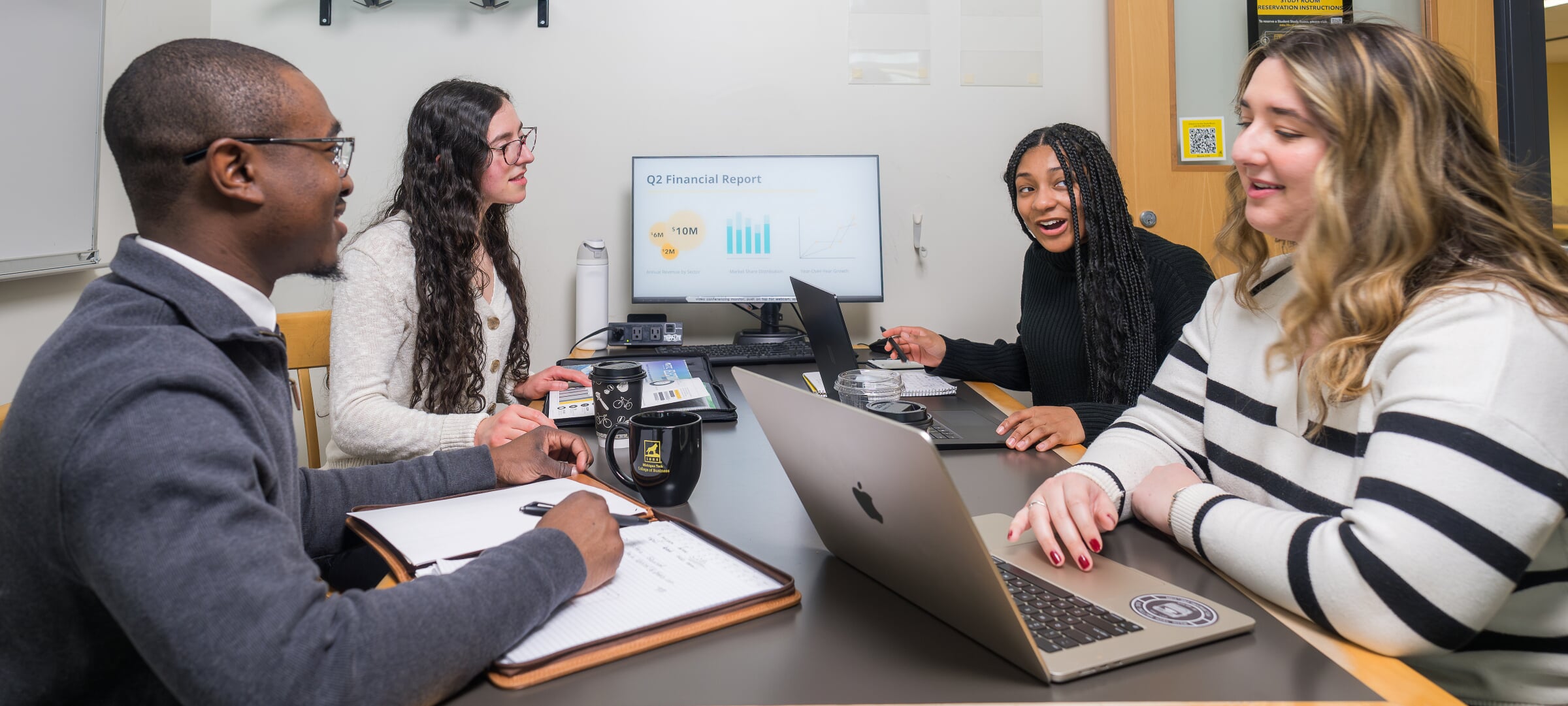 Four students sit around a table with notes and laptops, discussing a financial report.