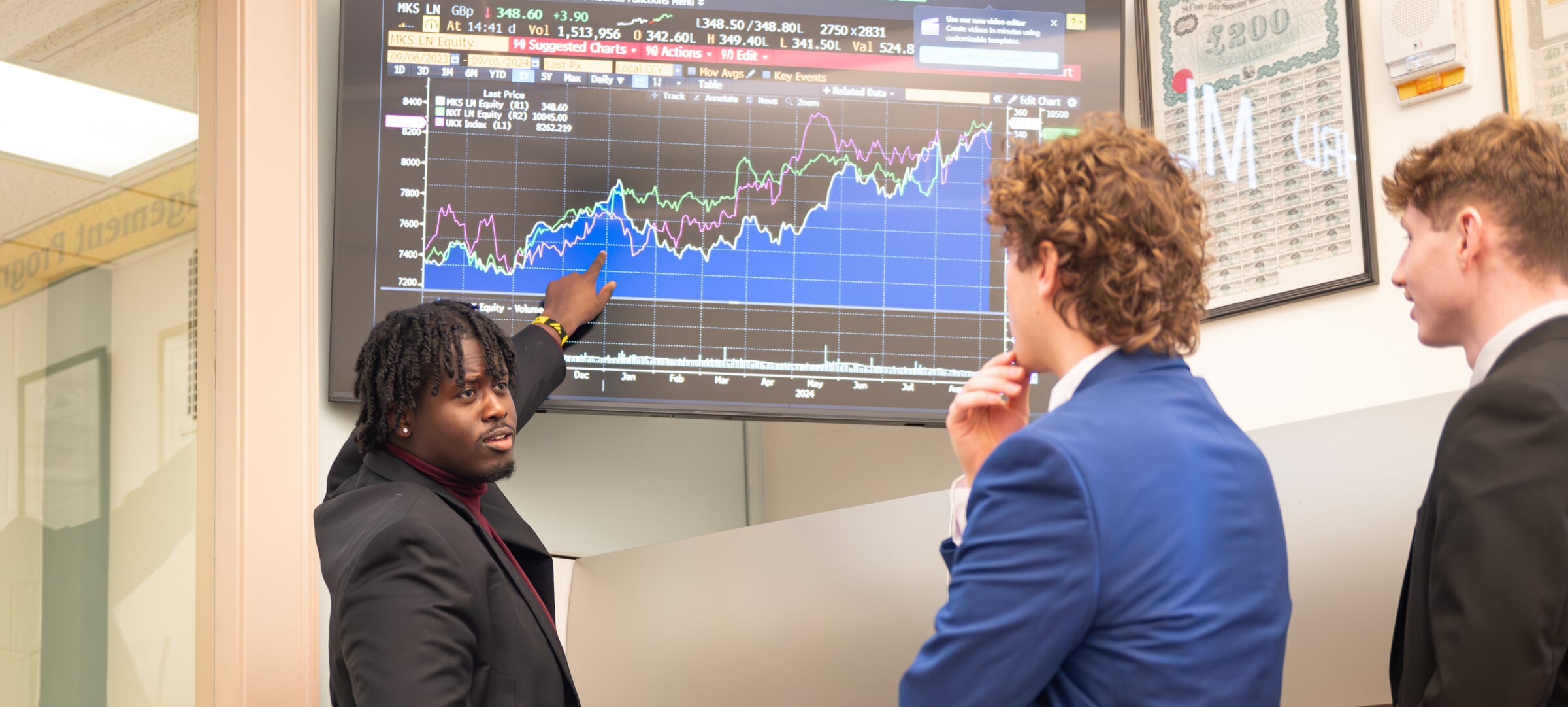 Three students discussing a financial report displayed on a TV.