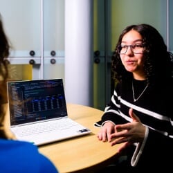 A student discusses the AI code seen on a laptop to another student, just out of frame.