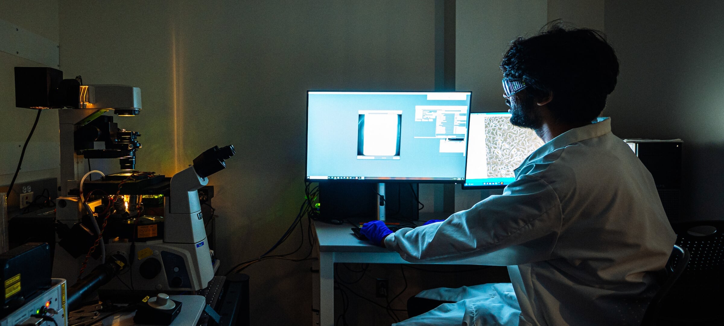 Scientific researcher inspects charts and data on two computer screens in a dim lab.