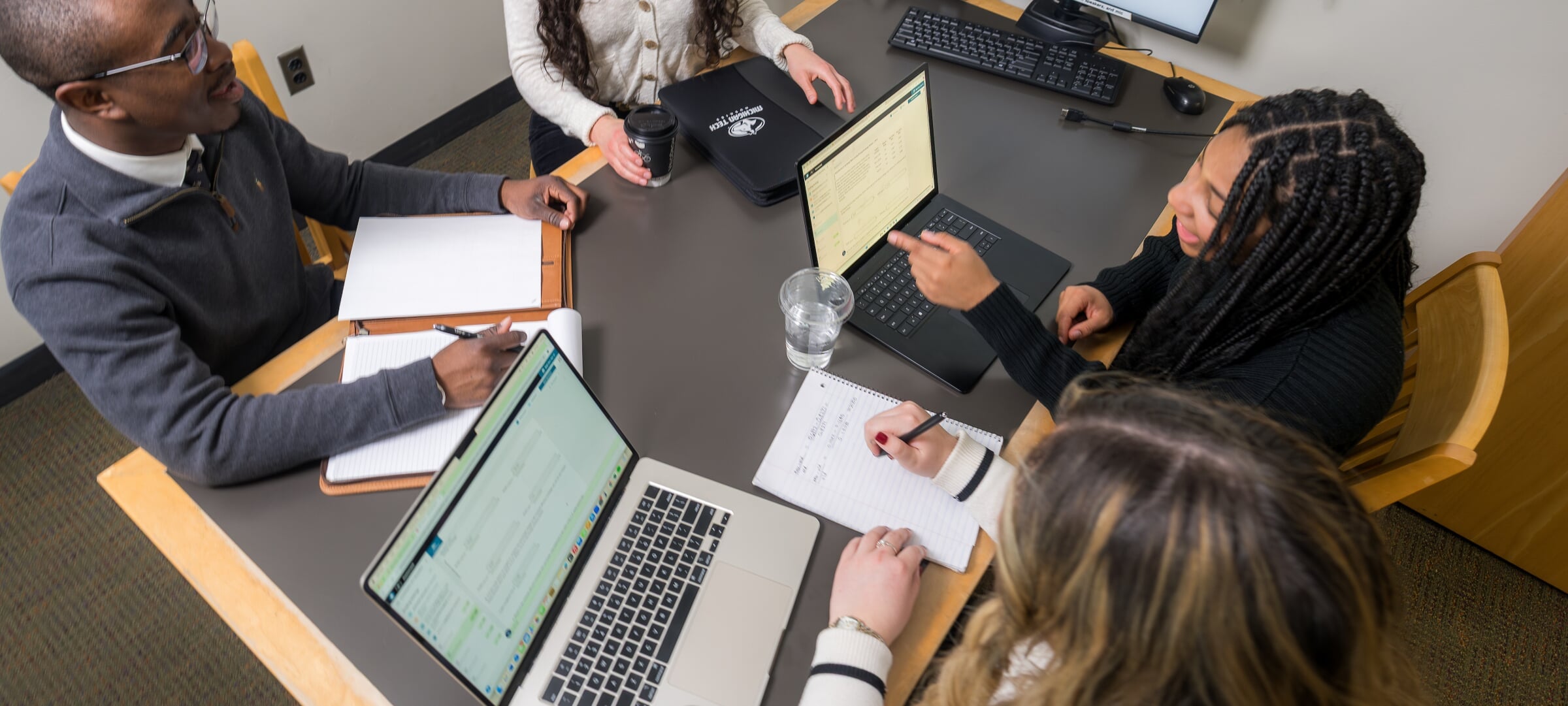 A group of business people talking and and taking notes on information from a pair of laptops.