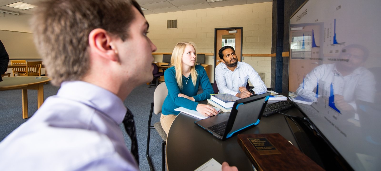 Graduate students sitting at a table with a large computer screen and laptop looking at statistical data.