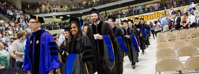 grad students in black robes with bright blue hoods on arms waling into commencement to get hooded. Lots of excitement on faces. 