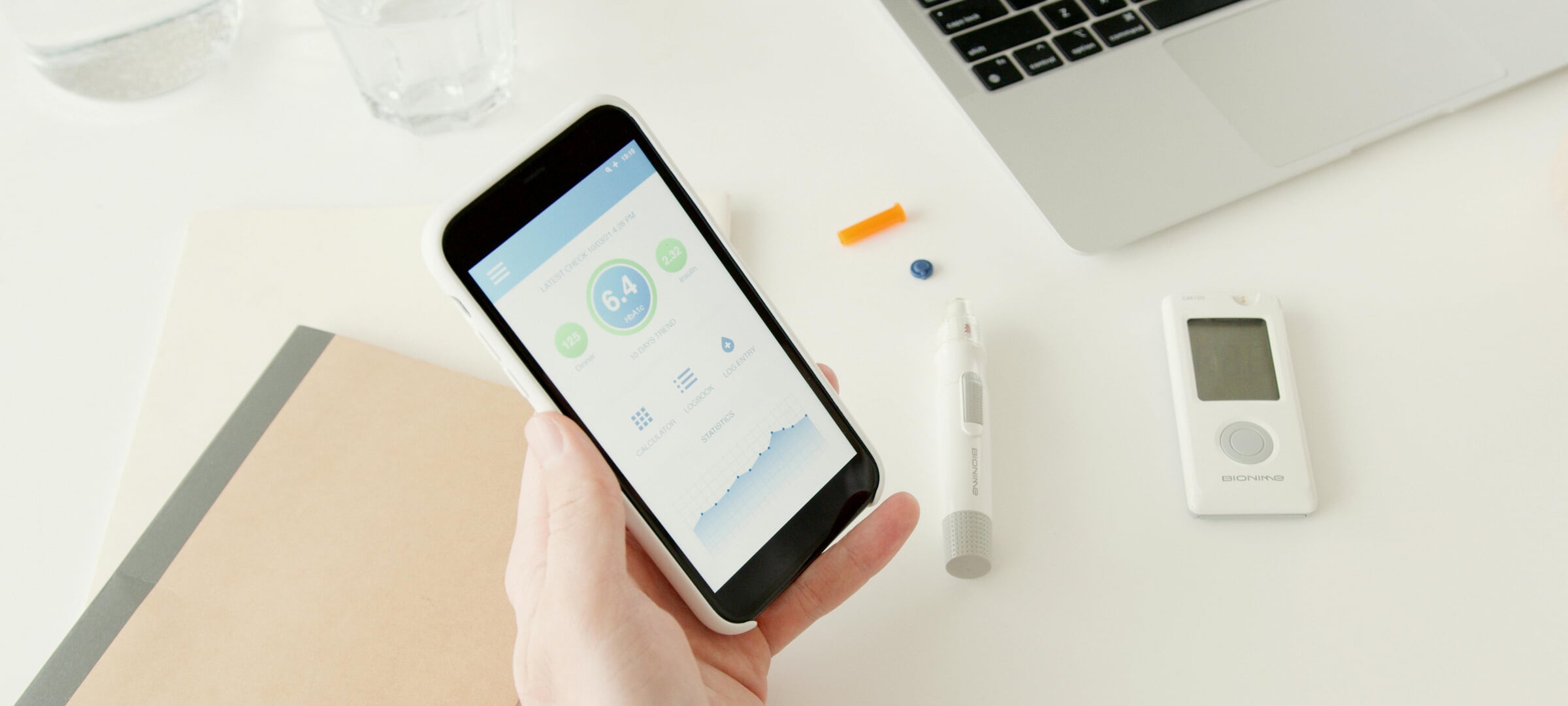 person holding a smartphone with a glucose reading on it while a glucose monitor and a computer are in the background