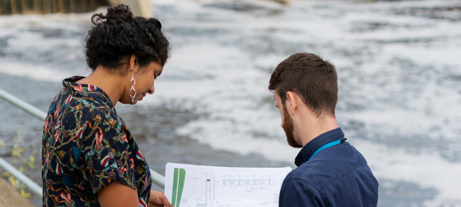 Two engineers looking at a plan while standing near a body of water.