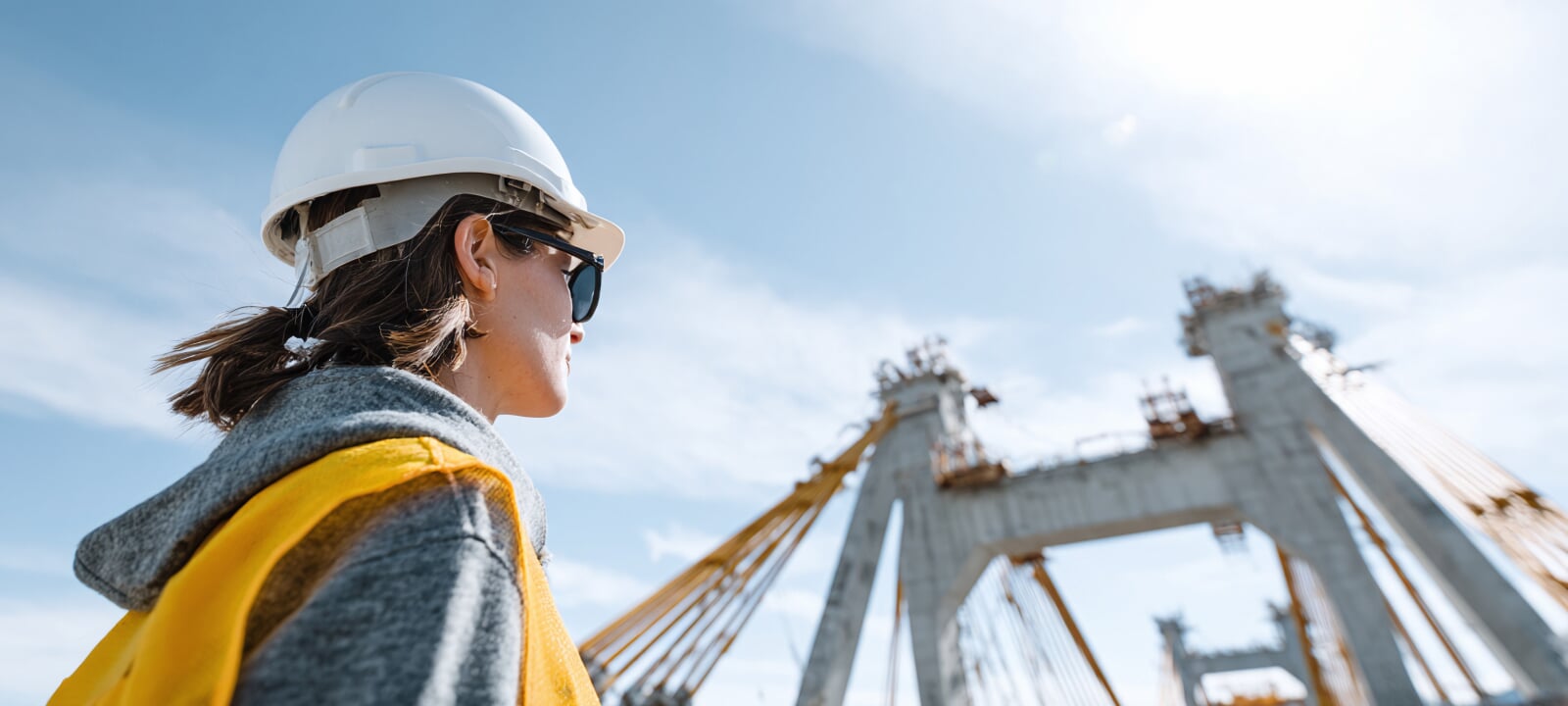 Female engineer wearing a hard hat and looking at a steel bridge, in the distance, which is under construction.