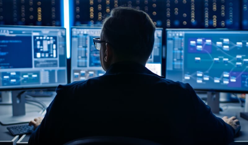 Maure cybersecurity professional looking at three computer monitors.