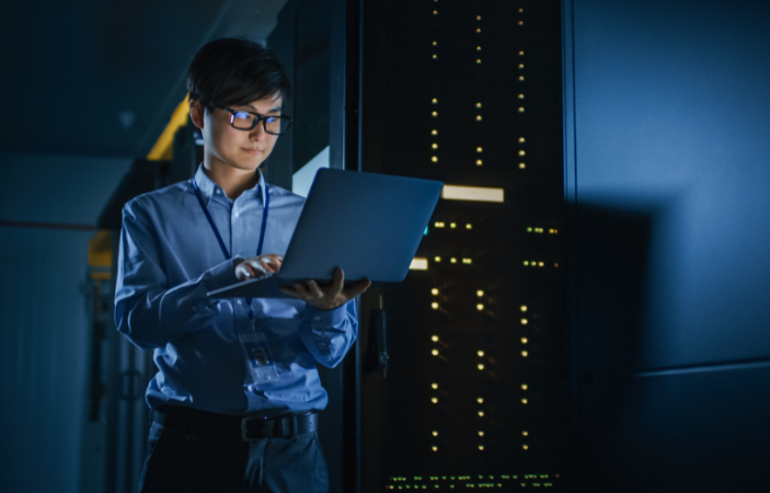 A young cybesecurity professional stands in front of a main frame computer.