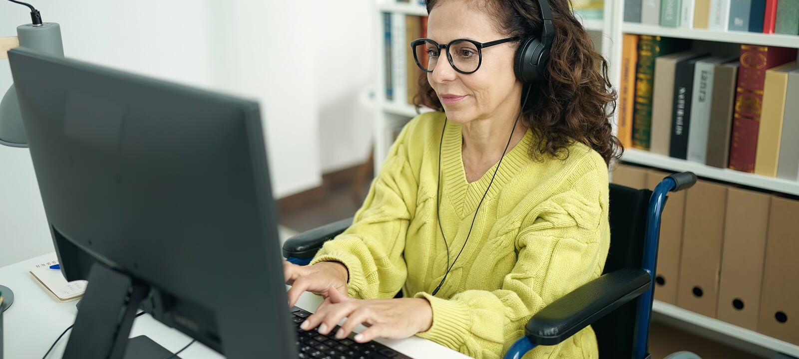 Older female Hispanic student wearing a yellow sweater while sitting in a wheelchair types at her computer while sitting in an office.