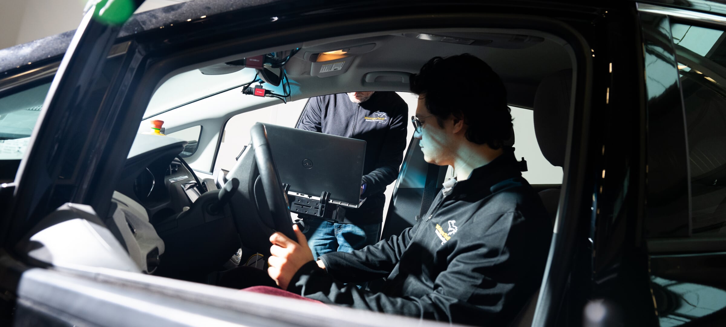 Grad student sits behind the wheel of a car while another researcher types on a laptop hooked into the car's systems.