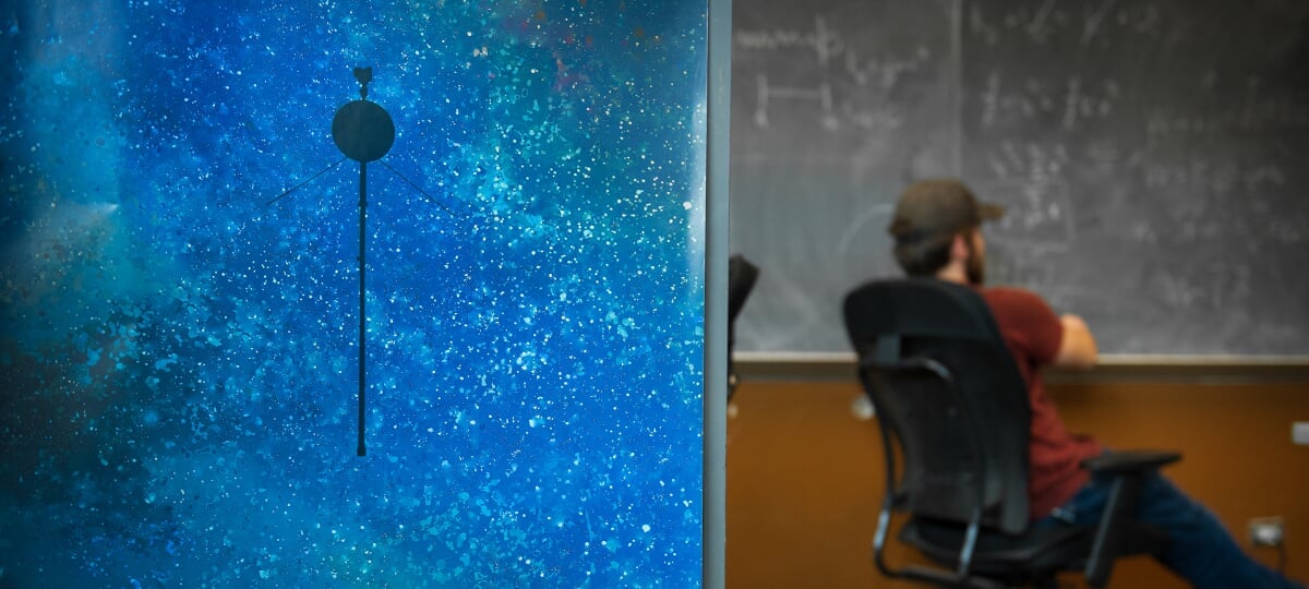 Closeup of a galaxy and Voyager probe laptop cover. A student sits by a chalkboard covered in equations.