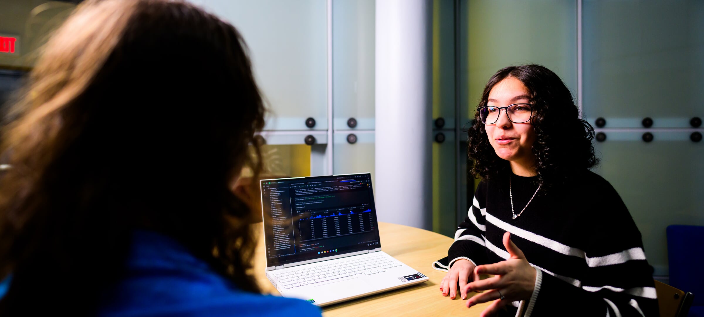 Two students discuss AI code, visible on the laptop on the table between them.