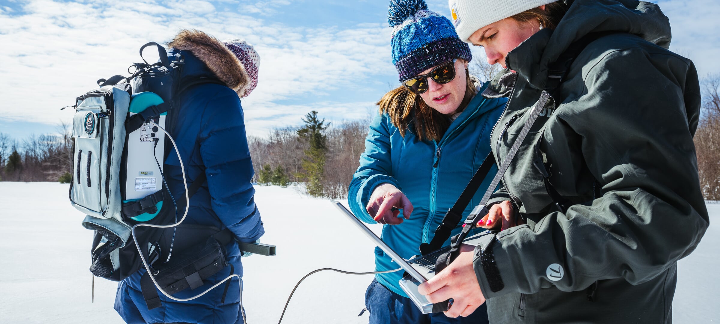 Three students using surveying equipment in a snowy field, inspecting the readings on a laptop.