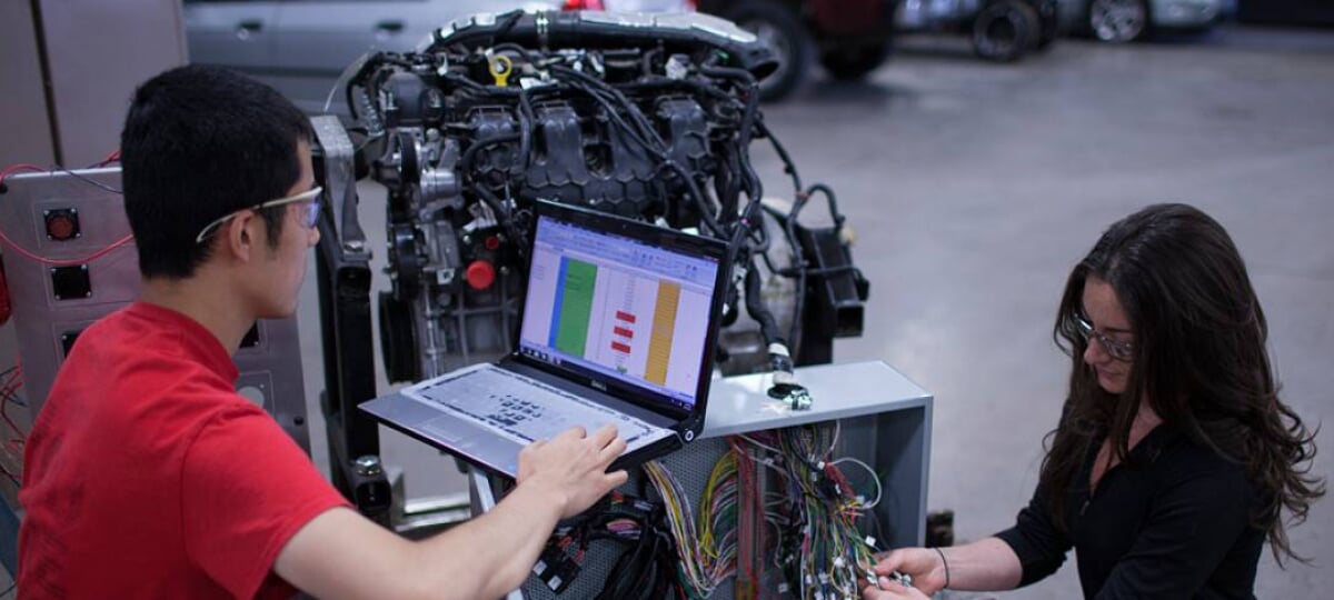 Two students, one examining laptop readouts from a combustion engine, one working on the wiring hooked up to the engine.