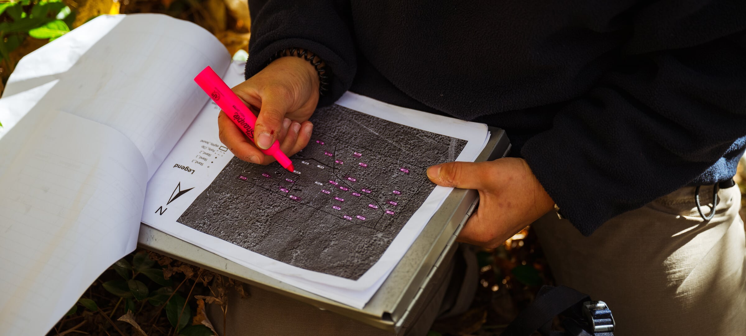 Student sitting in the forest, crossing off areas on a map with pink highlighter.