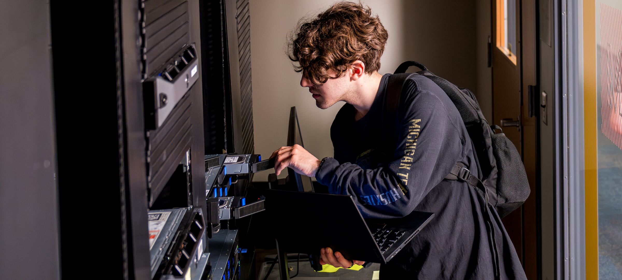 A student connects a laptop in their arms to a bank of servers.