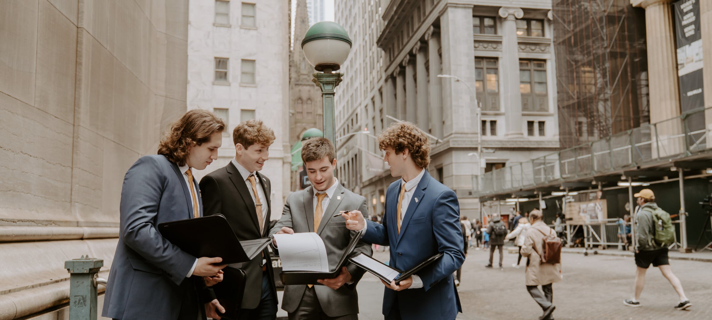 Four students in suits discuss information from portfolios beside a metropolitan street.