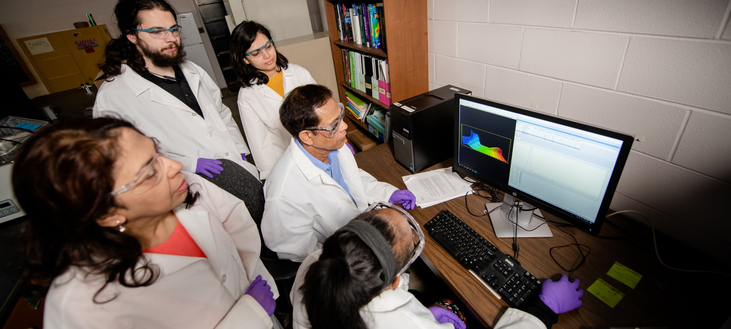 Grad students and faculty looking at a computer with labcoats, gloves and glasses