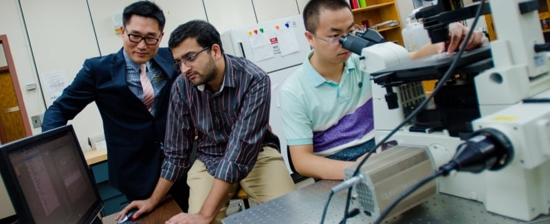 Professor, in suit and tie, is coaching two students who are capturing images from a microscope.