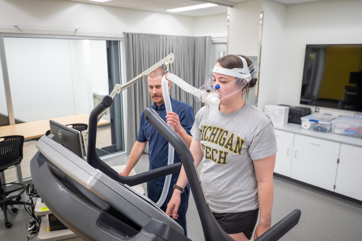 Student on treadmill with breathing apparatus, researcher looking on.