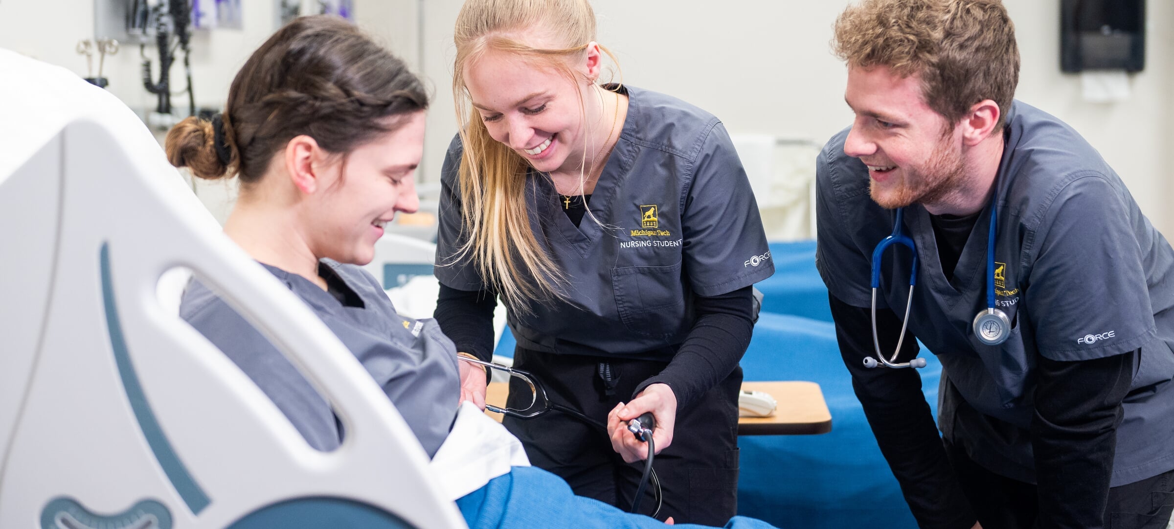 Three nursing students in a lab. Two are checking the blood pressure of a third.
