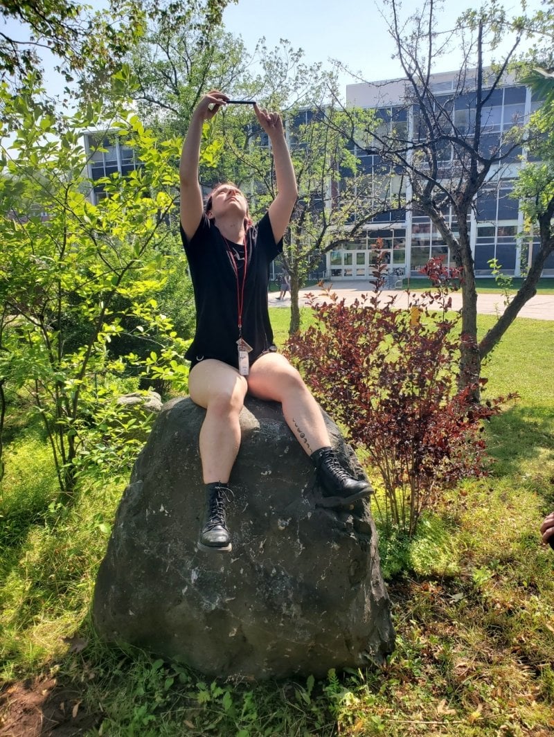 An image of a student sitting on a rock looking up into the ski at their phone.