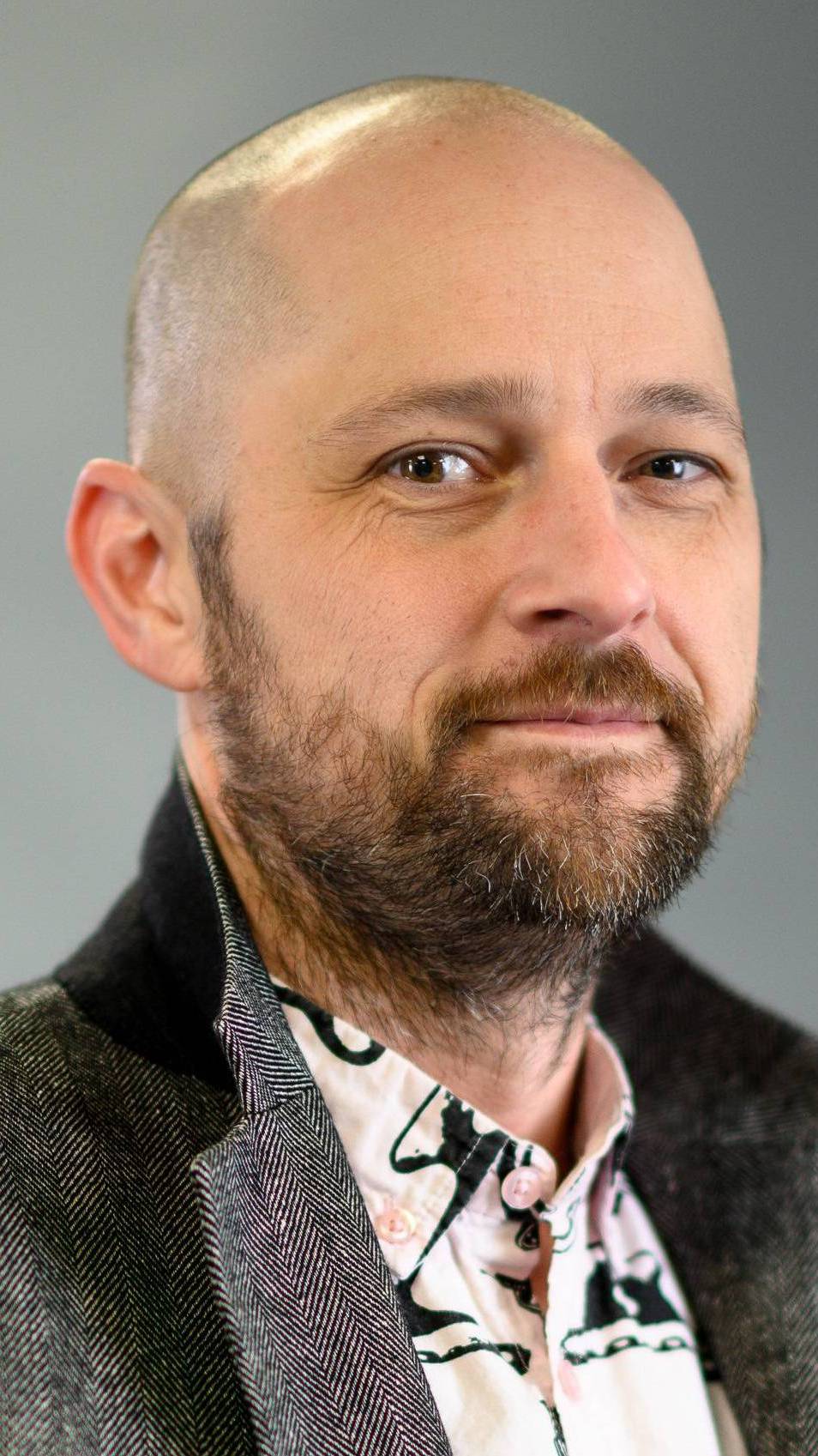 Headshot of man with shaved head, beard, pink button up shirt and blazer against a grey background