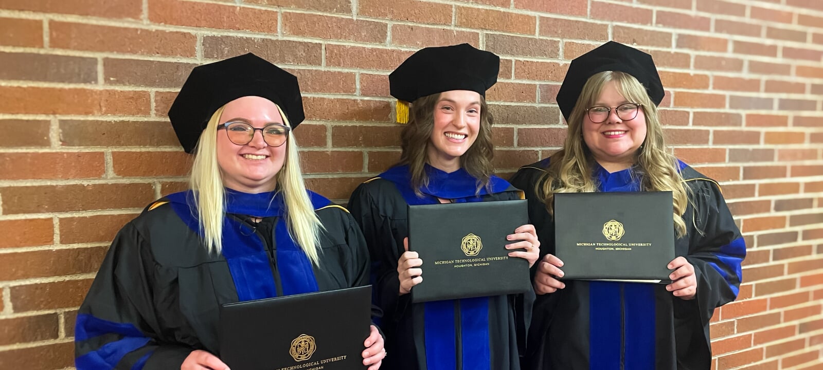 RTC PhD graduates Kendall Belopavlovich, Elizabeth Novotny, and Brilynn Janckila stand in their commencement regalia with diplomas against a brick wall.