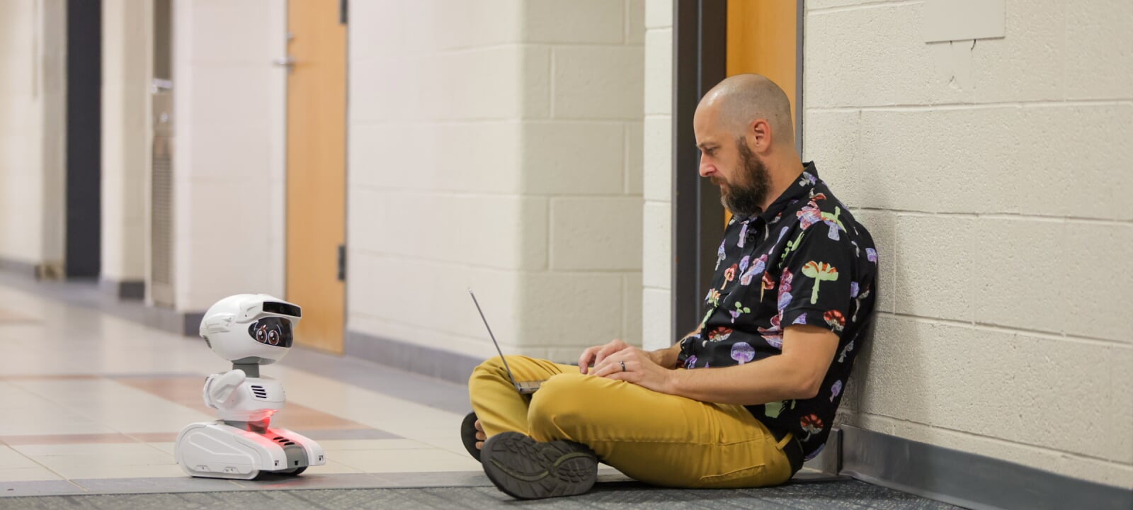 Dr. Jason Archer sits against a white brick wall in a hallway with his laptop while operating a small mobile robot that sits on the floor in front of him.