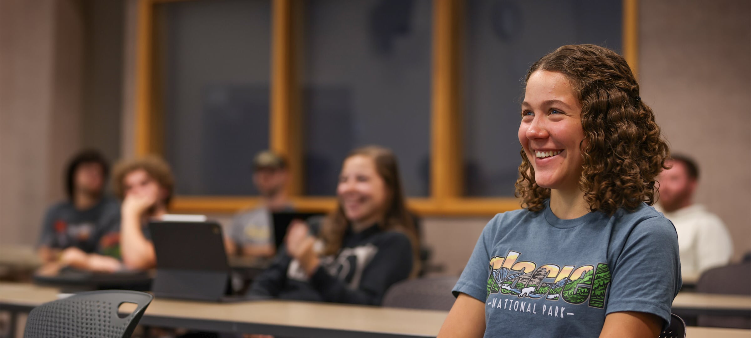 Several students in rows of tables with one in focus in the foreground, smiling.