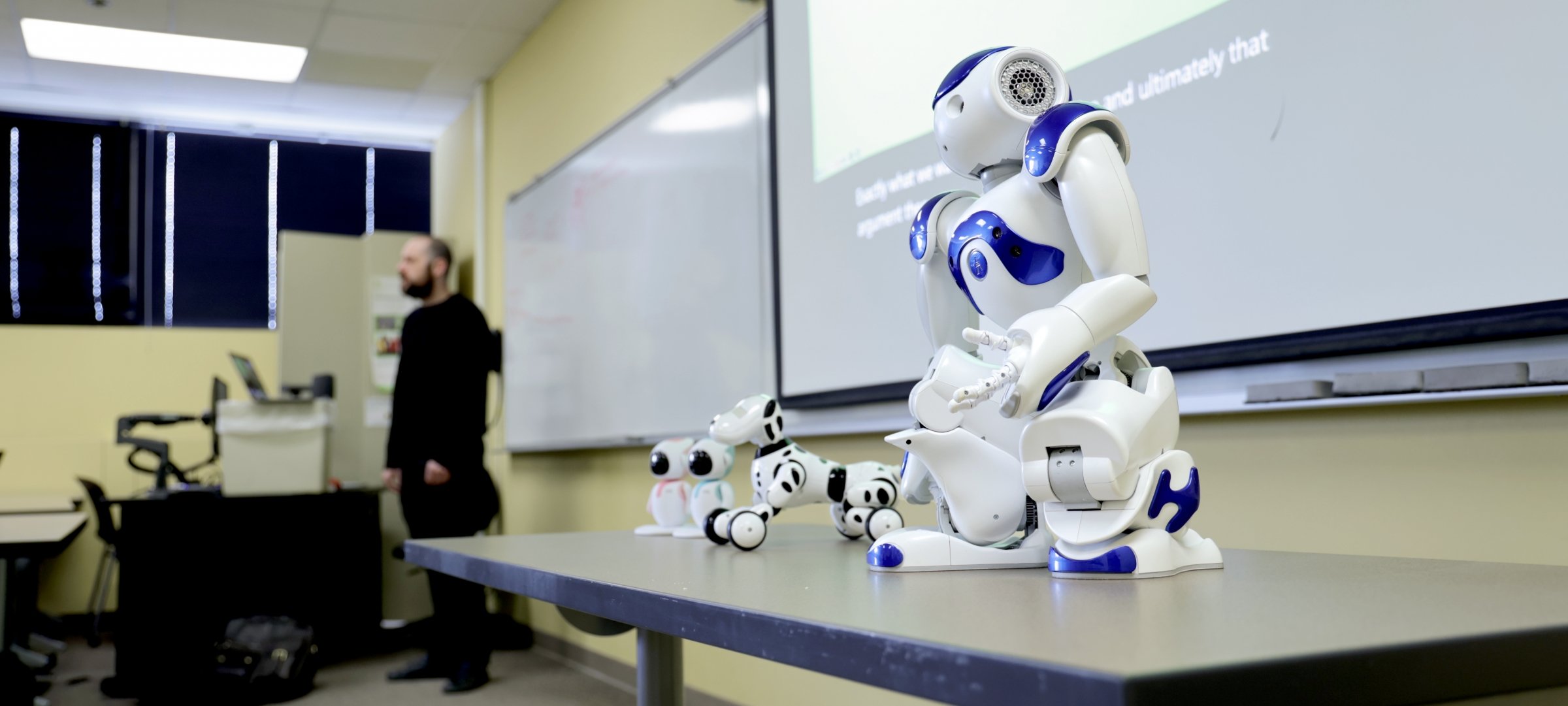 Four robots on a table in a classroom as a professor lectures in the background.