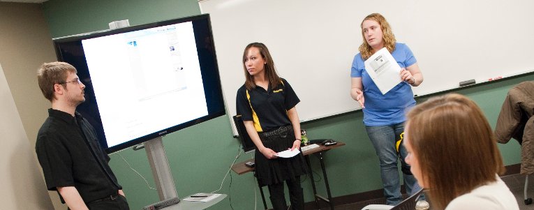 HDMZ Students standing inside a smaller media room discussing in front of a large monitor.