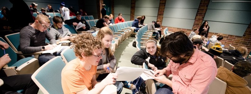 Faculty kneeling down to work with students in a lecture hall in the MonsterComp class.