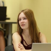A female student focuses on the instructor, out of view, with a male student out of focus in the foreground.