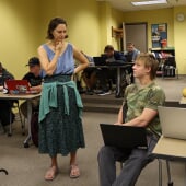 Wide-angle shot of instructor Claire Helakoski stands in the center of a classroom speaking to a student as the class works at their laptops around her.