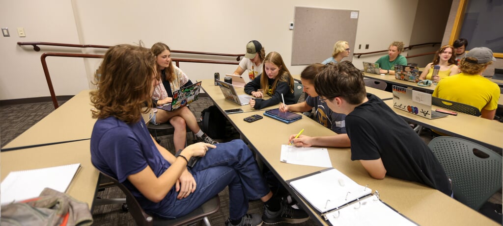 A group of students gather around a table with notebooks and laptops in group discussion.
