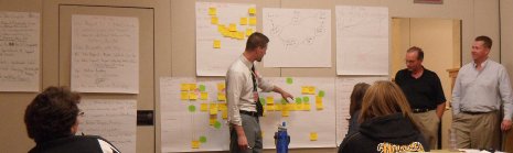 A man gestures to many post-it notes stuck on a wall, in front of a group of people.