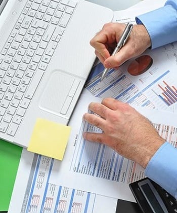 Hands holding a pen poised over papers in front of a laptop.