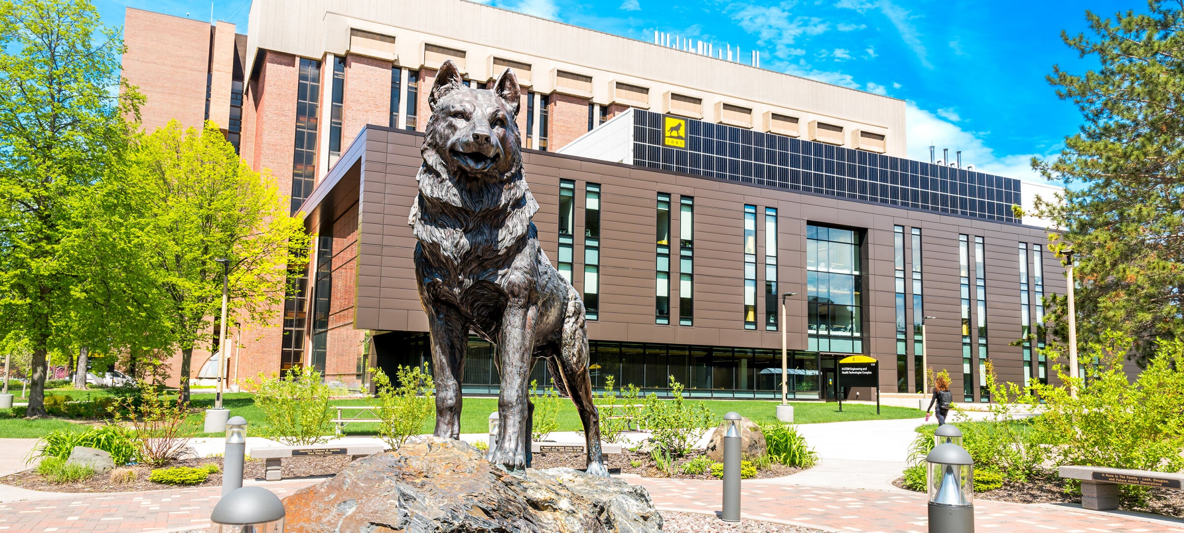 A springtime Michigan Tech campus featuring the Husky statue with the H-STEM and MEEM buildings in the background.