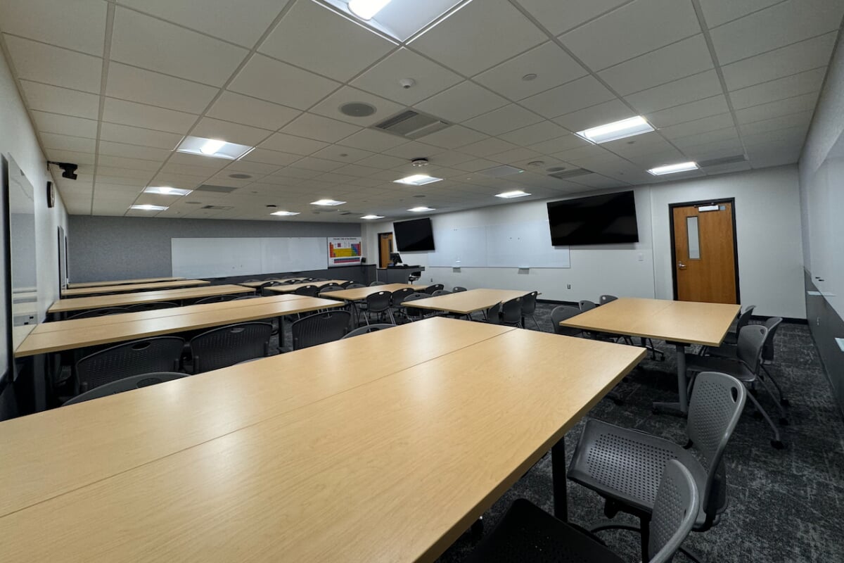 Wide, side-angle view from the back-left corner of the collaborative seminar room, showcasing the rows of student workstations, perimeter whiteboards, wall-mounted display monitors, and the main exit door.
