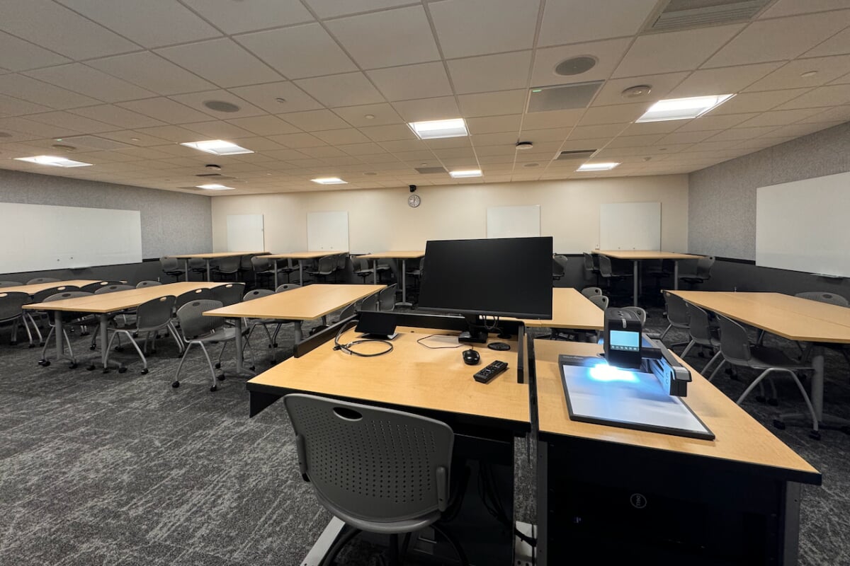 A collaborative seminar classroom layout featuring multiple modular rectangular student desks paired with grey plastic rolling chairs on a patterned grey carpet. The technology configuration includes a prominent instructor's media console in the foreground equipped with a desktop monitor and digital document camera, multiple flat-screen wall displays, and numerous independent whiteboards mounted along all perimeter walls for group collaboration.