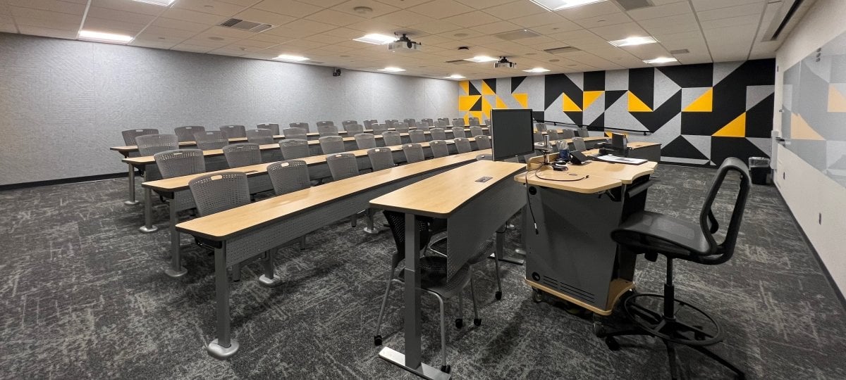 A lecture room layout viewed from the front instructional stage. Multiple ascending rows of long wooden student desks with mesh office chairs curve around the room on patterned grey carpet. The instructor's station features a wooden multimedia podium with a tall black drafting stool, a side computer monitor, and a prominent grey, black, and yellow geometric patterned accent wall in the background.