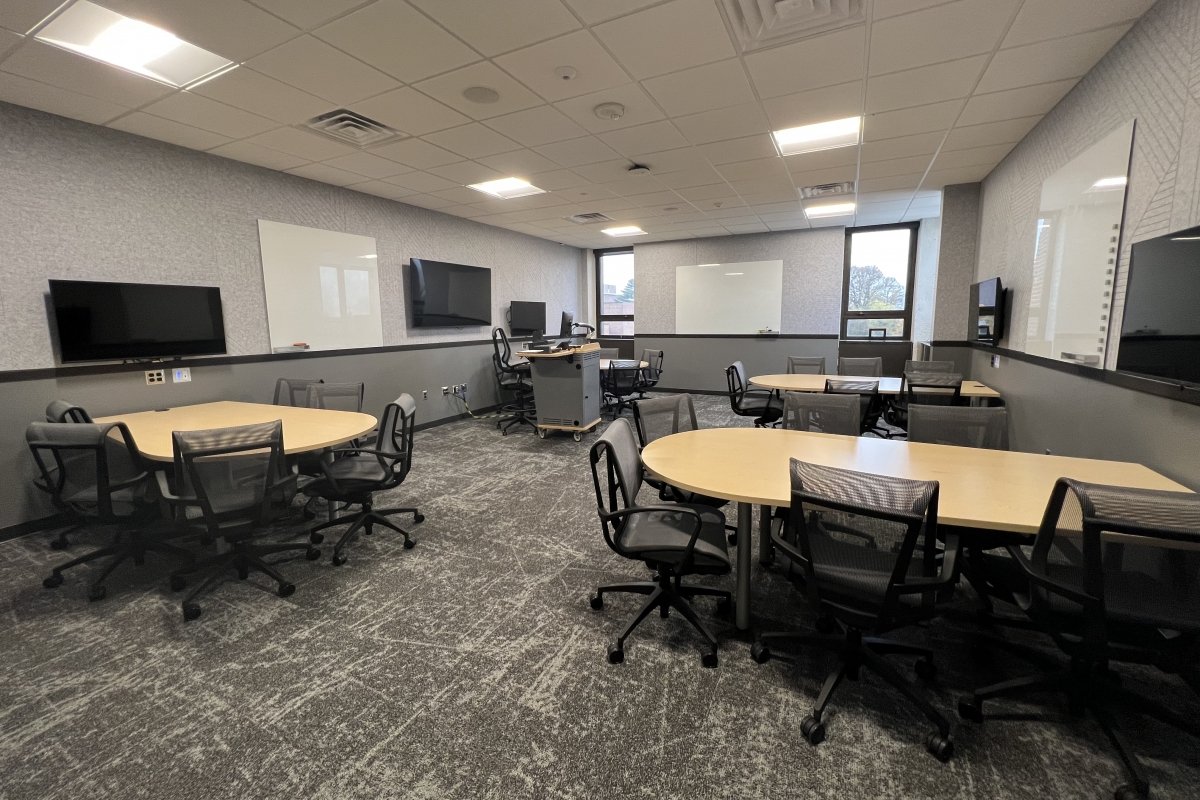 • An active learning classroom layout featuring multiple large, oval collaborative team tables paired with black mesh rolling chairs on a patterned grey carpet. The room's instructional technology configuration includes multiple flat-screen digital displays and whiteboards mounted around the perimeter walls for group work, with a central instructor's media podium positioned near the back.