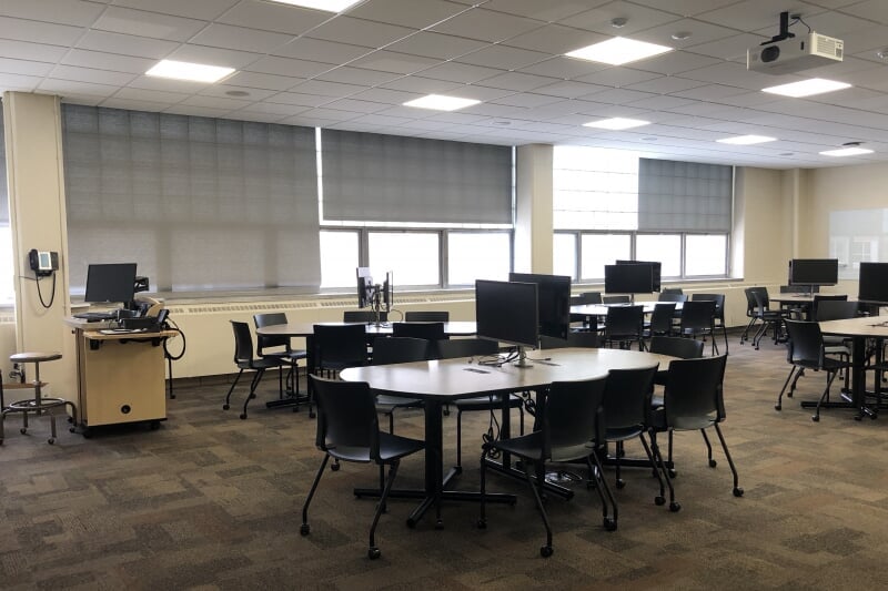 Alt Text: An active learning classroom layout featuring multiple large, oval collaborative team tables paired with black mesh rolling chairs on a patterned grey carpet. The room's instructional technology configuration includes multiple flat-screen digital displays and whiteboards mounted around the perimeter walls for group work, with a central instructor's media podium positioned near the back.