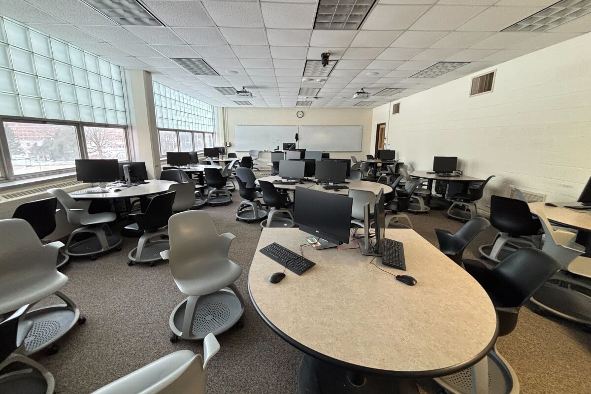A collaborative computer lab classroom layout featuring multiple teardrop-shaped team tables arranged throughout the room. Each table workstation is equipped with dual desktop computer monitors, keyboards, and mice, paired with grey and black mobile caster chairs featuring under-seat storage trays. The room technology includes ceiling-mounted digital projectors pointing toward a large whiteboard at the front, with large glass-block perimeter windows providing natural light along the side wall.